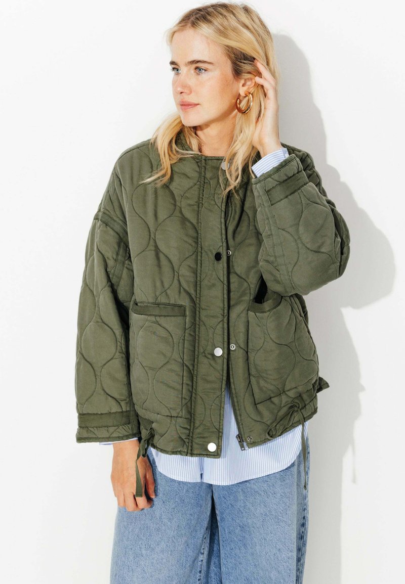 Veste matelassée vert olive avec un col rond, fermeture à pression et deux poches latérales, portée par-dessus une chemise rayée bleu et blanc.