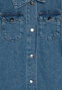 Jeans košulja u svijetloplavoj boji s dva džepa na prsima, srebrnim dugmadi na kopčanje i ovratnikom. Tkanina ima blago teksturirani izgled.