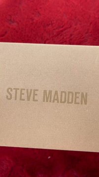 Pruun pappkast, mille peal on suurtähtedega kirjas "STEVE MADDEN", asetatud punasele tekstureeritud pinnale.