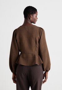 Hnědý pruhovaný top s peplum střihem a dlouhými, vypufovanými rukávy, zaobleným výstřihem a přiléhavým pasem. Materiál má hladkou strukturu. V kombinaci s tmavými kalhotami.