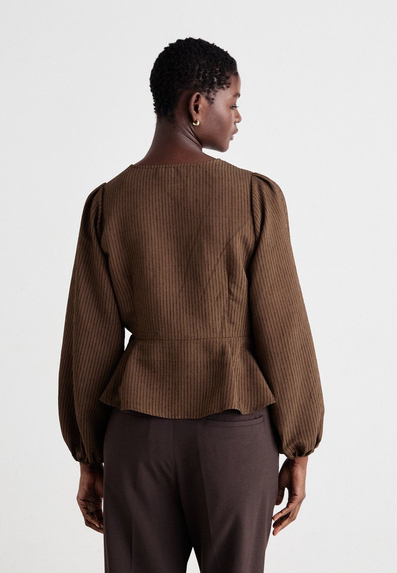 Brązowa, pasiasta bluzka peplum z długimi, bufiastymi rękawami, okrągłym dekoltem i dopasowaną talią. Materiał ma gładką teksturę. W zestawie z ciemnymi spodniami.