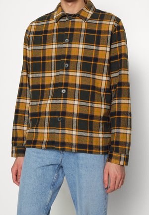 Mężczyzna w koszuli na guziki z długim rękawem o kraciastym wzorze w kolorach brązowym, czarnym, białym i pomarańczowym oraz w jasnoniebieskich dżinsach, stojący na białym tle.