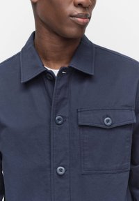 Marinblå arbetsjacka i slitstark tyg med krage, två knappar och en bröstficka. Har en texturerad yta och matchande knappar.