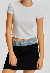 Gekürztes weißes Baumwoll-T-Shirt kombiniert mit einem schwarzen Rock, der einen hellen Denim-Taillenbund hat. Einfaches Design mit kurzen Ärmeln und Rundhalsausschnitt.