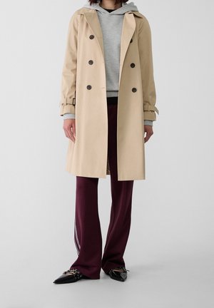 Personne portant un trench beige par-dessus un sweat à capuche gris, associé à un pantalon bordeaux avec des rayures blanches et des chaussures pointues noires.