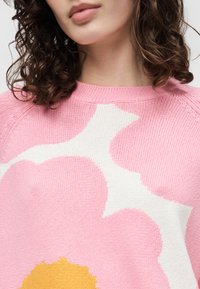 Γυναίκα με σγουρά σκούρα μαλλιά που φοράει ένα ροζ πλεκτό πουλόβερ με μεγάλο λευκό και κίτρινο σχέδιο λουλουδιού στο μπροστινό μέρος.