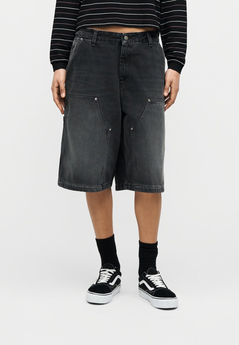 Person trägt schwarze, übergroße Jeansshorts mit großen Fronttaschen, schwarz-weiße Turnschuhe und schwarze Socken.