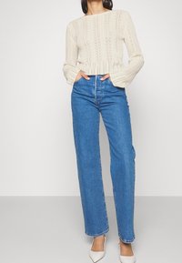 Cremfarbener gestrickter Pullover mit einem gerippten Muster, kombiniert mit hochgeschnittenen blauen Straight-Leg-Denim-Jeans und spitzen weißen Absatzschuhen.