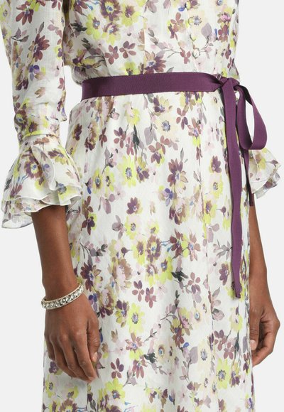 Ea poartă o rochie albă cu flori galbene și mov, o curea violet legată în talie și o brățară argintie strălucitoare la încheietura mâinii.