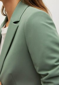 Jasnozielony żakiet o strukturalnym kroju, z klapami w kształcie litery V i gładkim, teksturowanym materiałem, widoczny od ramienia w dół.