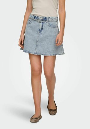 Donna che indossa una minigonna in denim azzurro chiaro con una cintura a doppia fibbia e ballerine marroni, in piedi davanti a uno sfondo semplice.