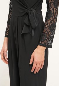 Vêtement noir avec manches longues en dentelle, détail noué à la taille et tissu fluide, montré sur le torse et les bras d'une personne.