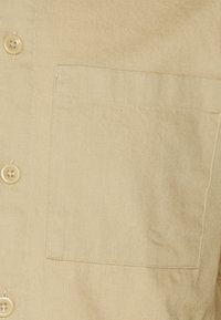 Beige Hemd mit Knopfleiste aus strukturiertem Stoff, ausgestattet mit einer einzelnen Brusttasche und runden Knöpfen zum Verschließen.