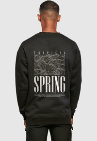 Mustaline särk, millel on valge graafiline disain, millel on tekst "TROPICAL SPRING" ja lehe mustrid seljal. Pehme tekstuur, ümmargune kaelus.