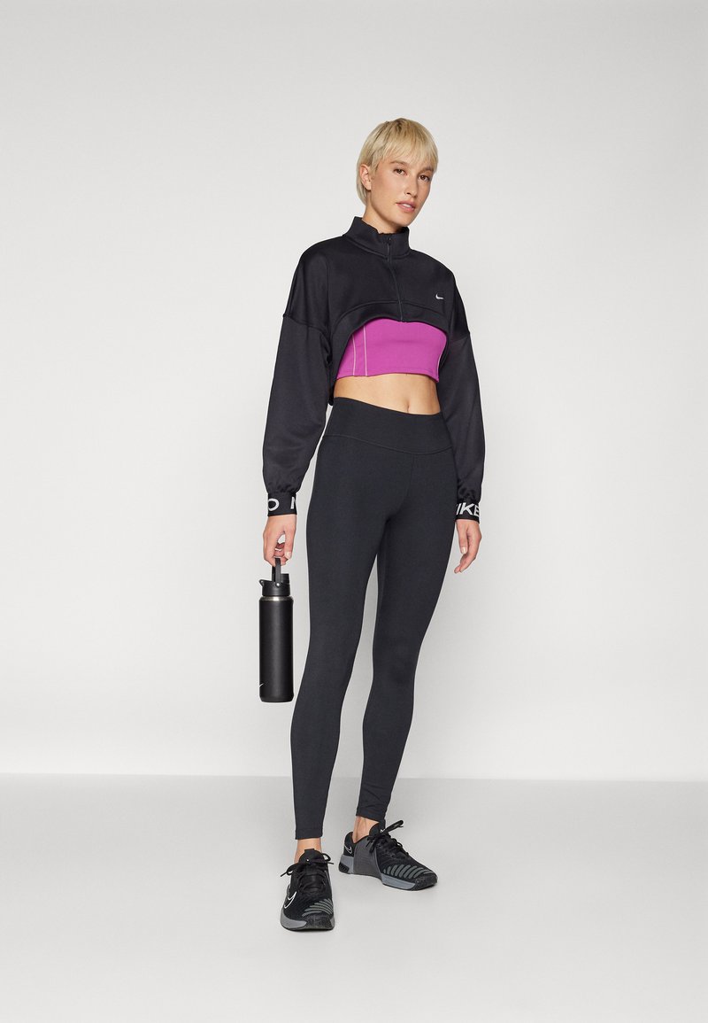 Schwarze, kurze Zip-Jacke mit lila Panele, schwarze hochgeschnittene Leggings und schwarze Sportschuhe. Hält eine schwarze Wasserflasche.