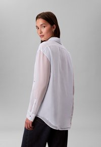 Blouse blanche avec manches transparentes, col classique, poignets boutonnés et ourlet légèrement arrondi. Modèle légèrement tourné vers l'arrière, mettant en valeur le design.