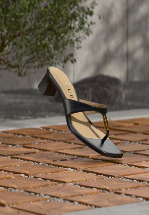 Zwarte sandaal met hak, teenlus en gouden accent, geplaatst op een houten en stenen geplaveid oppervlak buiten nabij groen.