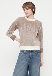 Kvinde med kort brunt hår iført en beige og hvid stribet striktrøje og løse blå jeans, som poserer med den ene hånd på hoften.