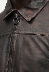 Κοντινή λήψη στο γιακά από σκούρο καφέ δερμάτινο μπουφάν και κατακόρυφη φερμουάρ με στρογγυλό τράβηγμα, που δείχνει την υφή του δέρματος και τις λεπτομέρειες των ραφών.