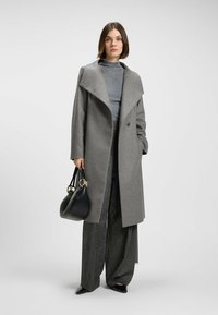 Cappotto grigio in misto lana con ampio colletto, doppi bottoni e tasche laterali, abbinato a un top grigio e una borsa nera, indossato con pantaloni a gamba larga.