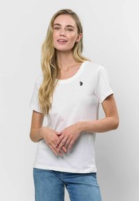 Balta kokvilnas T-kreklā ar apaļu izgriezumu un īsām piedurknēm, ar mazu melnu izšuvumu uz krūtīm. Standarta piegriezums.