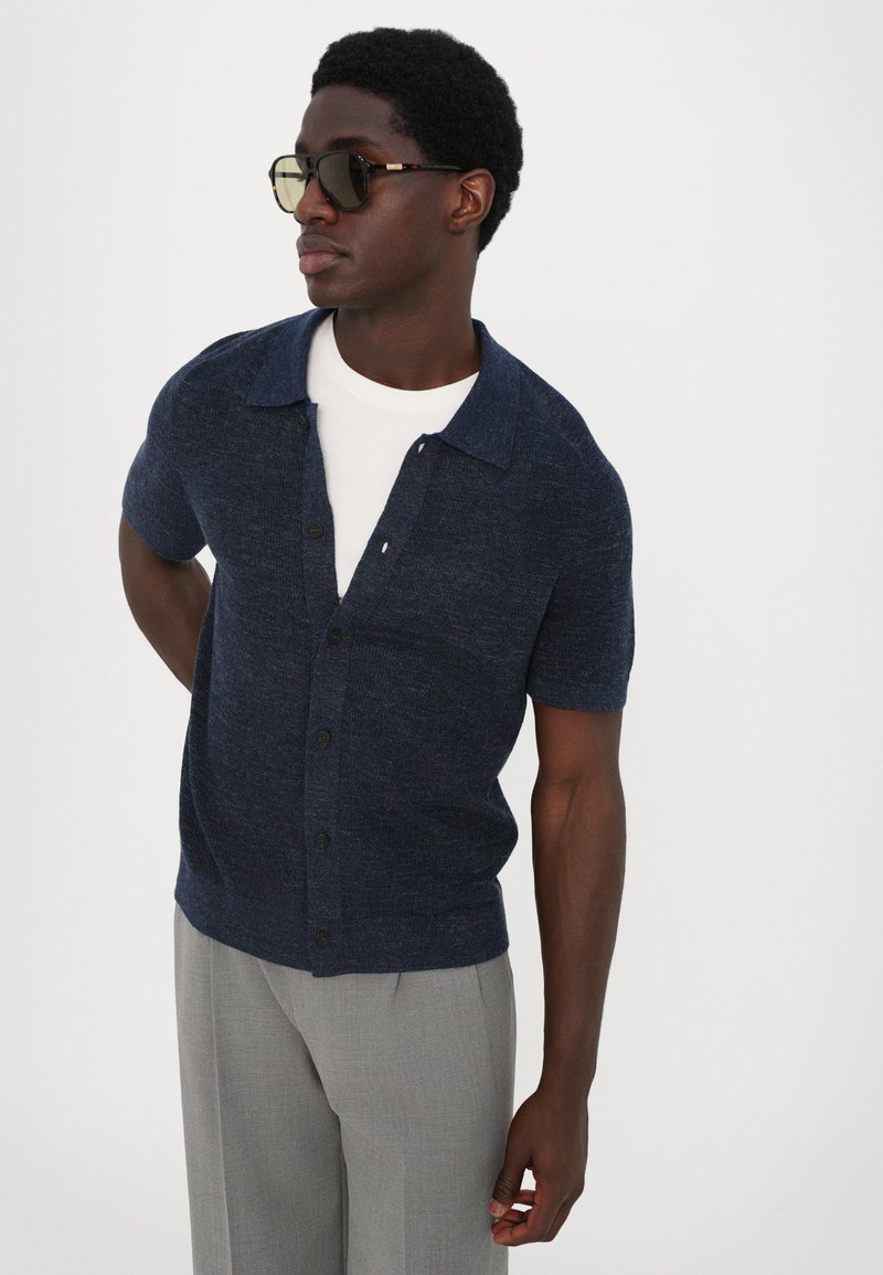 Navy short-sleeve knitted cardigan with a button-down front, featuring a spread collar. Worn over a white shirt, paired with gray trousers.