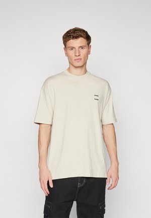Jeune homme aux cheveux courts et bouclés, portant un t-shirt beige ample et un pantalon cargo noir, debout devant un fond blanc uni.