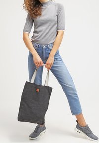 Grå canvas tote-väska med ljusgrå remmar, rektangulär form och en liten brun läderetikett. Modell som håller väskan, ihop med denim.