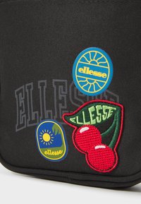 Borsa di stoffa nera con toppe circolari blu e verdi con loghi e un design texturizzato di ciliegie rosse. Gli accenti includono dettagli ricamati.