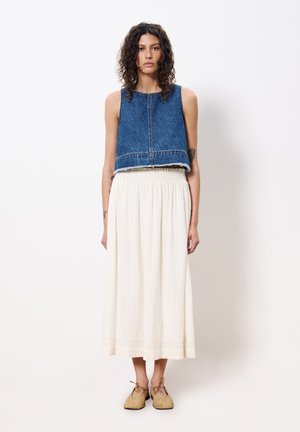 Denim-Crop-Top mit fransigem Saum, kombiniert mit einem fließenden, cremefarbenen, plissierten Maxirock. Das Model trägt tanfarbene Schnürschuhe.