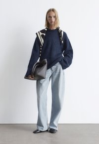 Marineblauen, flauschigen Pullover, hellblaue weite Jeans, schwarze flache Schuhe und eine dunkelgraue Tasche. Schwarz-weiß gemusterter Schal über den Schultern drapiert.