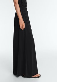 Femme portant une longue jupe noire et des sandales noires, debout de profil sur un fond clair uni.