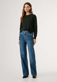 Pull en maille noir à manches longues, associé à un jean bleu taille haute avec un motif subtil. Le mannequin porte des bottes noires à talons.