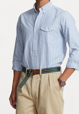 Homme portant une chemise bleu clair à rayures verticales, manches retroussées, pantalon beige, ceinture en tissu rayée et montre-bracelet.