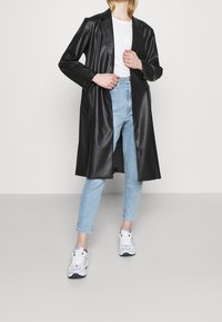 Svart fusk läder lång kappa med notched krage, i kombination med ljusblå denimjeans och vita sneakers. Slät textur, rak passform.
