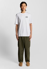 Witte katoenen t-shirt met een ronde hals en een zwarte graphic op de linker borst, gecombineerd met olijfgroene cargobroek en beige instap schoenen.