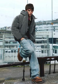 Grijze pufferjas, zwart gelaagd shirt, lichtblauwe geborduurde jeans en bruine dikke laarzen; met één voet op een bankje staand.