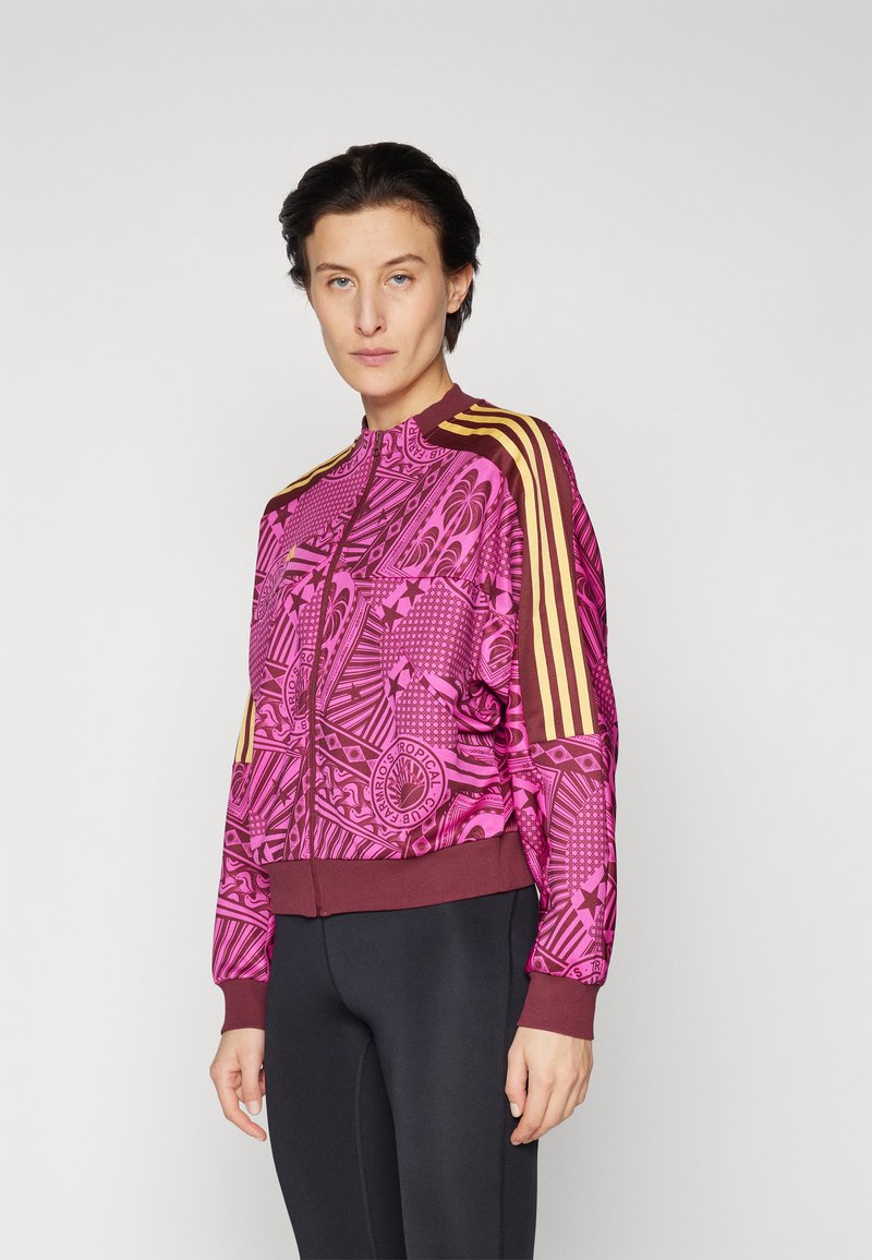 Trainenjack in levendig roze met ingewikkelde patronen. Beschikt over bruine boorden en een rits, plus drie gele strepen op de schouders.