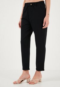 Pantalons de jogging en coton noir avec taille élastique, détails de boutons, poches latérales et jambes fuselées, associés à des sandales à lanières de couleur claire.