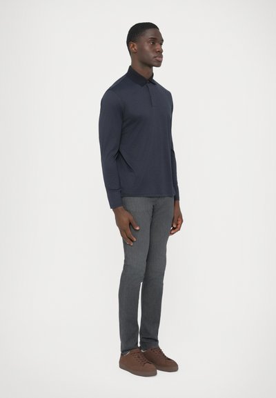 Tengerészkék hosszú ujjú galléros póló szürke, karcsúsított nadrággal és barna, velúr sportcipőkkel párosítva, fehér háttér előtt állva.