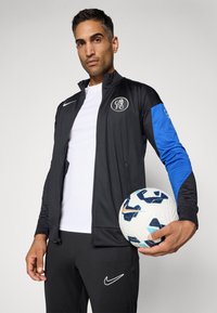 Black and blue athletic jacket with zip, logo on chest, white shirt underneath, black pants with logo, holding a patterned soccer ball.