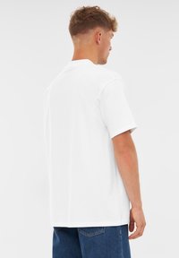 Vit kortärmad t-shirt i bomull med rund halsringning och normal passform, bärs ihop med blå denimjeans sett från en bakvinkel.