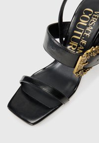 Black leather sandal with two wide straps, square toe, and a gold-tone decorative buckle. Interior features text: “Versace Jean Couture.”