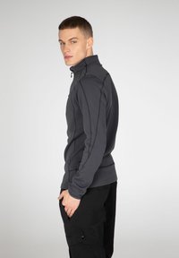 Tamnosivi zip-up sako s visokim ovratnikom. Ima šivane šavove, glatku teksturu i duge rukave. Model nosi crne hlače, ruke u džepovima.