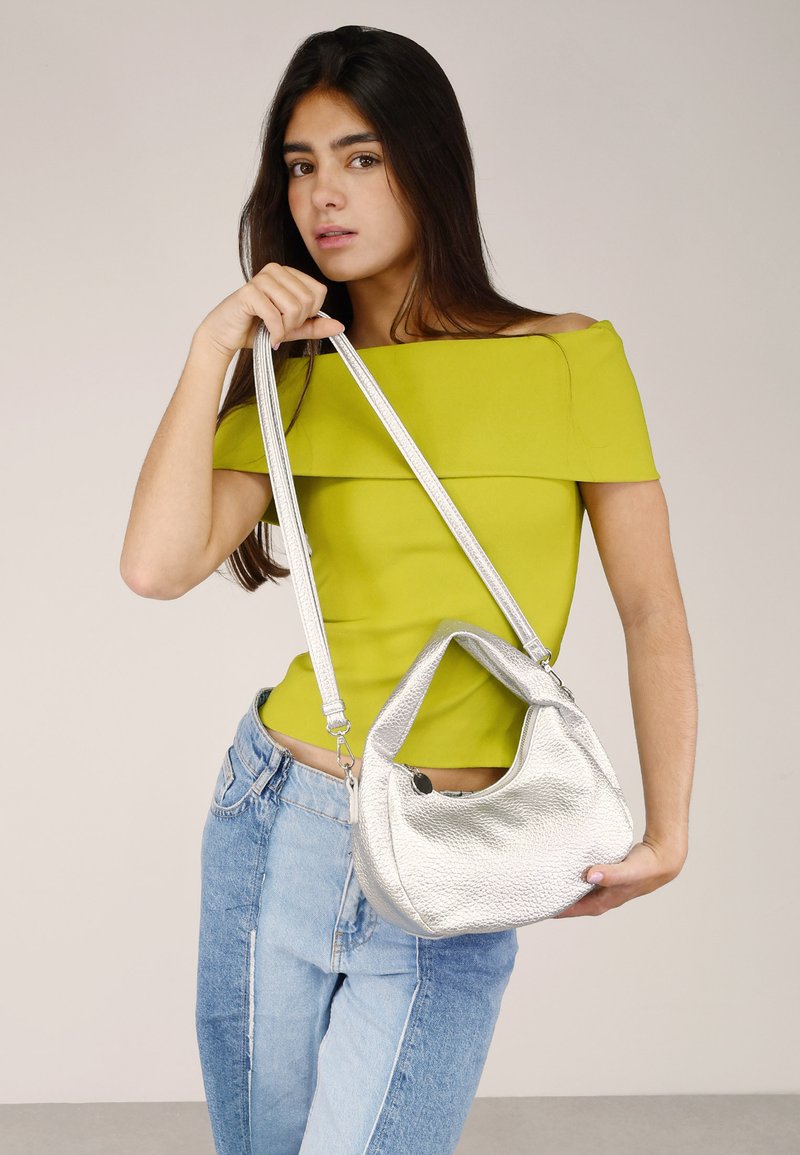 Bolso de mano de piel plateada con acabado texturizado, forma curvada y doble asa. La modelo lleva una blusa verde brillante sin mangas y jeans de denim.
