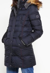 Marineblauwe donzen jas met een capuchon afgezet met bont, voorzien van een rits- en drukknoopsluiting, zijzakken en gestructureerde grijze accenten op de kraag.