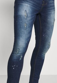 Denim jeans in dark blue with faded and distressed patches. Features light blue splatter accents and visible stitching along the seams.