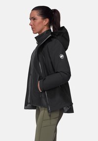 Svart huppjacka med dragkedjefickor, texturerad tyg och en cirkulär logotyp på vänster ärm; bärs över olivgröna leggings.