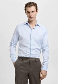 Svetlo modra srajca z ovratnikom v obliki konice, z dolgimi rokavi in sprednjim zapenjanjem na gumbe, kombinirana s temno rjavimi hlačami. Gladek tekstura tkanine.