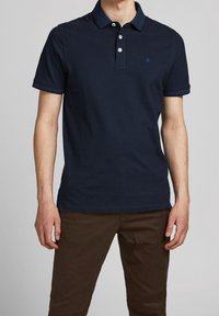 Marinblå piké av mjuk bomull, med tre knappar, korta ärmar och en subtil logotypaccent på bröstet.